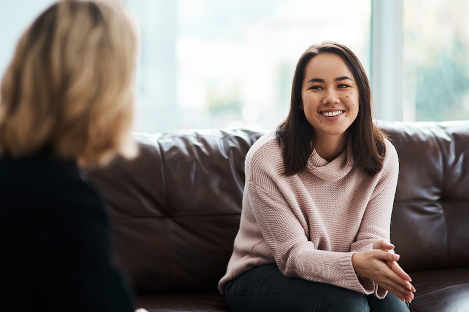 A view of two people talking. The camera is behind the person on the left with the person on the right sits on a couch and smiles.