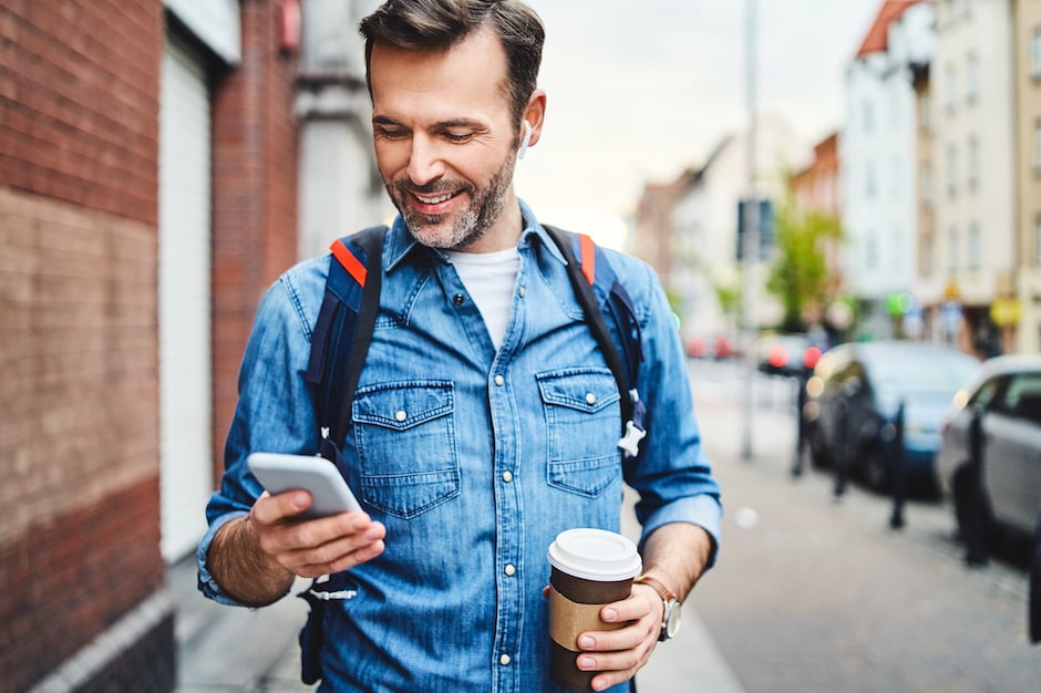 A man walks along a street looking at his mobile phone in his right hand, while carrying a coffee in his left hand. 